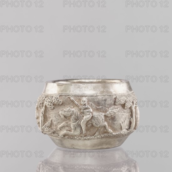 Bowl, South Asian culture, Indian culture, Indian art; hunting iconography: tigers, horses, elephants, galloping rider on horseback, warriors; plants; applied art; 19th/20th century; India; vessel; weight 155 g; height 5.4 cm; diameter 8.0 cm, A silver-colored rounded bowl sits against a light gray background, with a raised decorative band around its middle depicting a scene in bas-relief. The relief shows a figure seated on a horse with one arm raised, surrounded by textured foliage and vegetation motifs, including leafy forms and clusters suggesting trees. The bowl has a smooth upper rim and a slightly flared base, with a reflective, metallic surface showing subtle variations in sheen and patina in tones of silver and pale gray.
