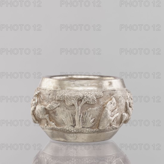 Bowl, South Asian culture, Indian culture, Indian art; hunting iconography with tigers, horses, elephants, galloping rider on horseback, warriors, and plants; applied art; 19th/20th century, India; vessel; weight 155 g; height 5.4 cm; diameter 8.0 cm, A small rounded metal vessel with a matte silver surface sits against a light gray background and reflects faintly below. The vessel features a raised decorative band around its middle with sculpted natural motifs including a stylized tree with textured canopy, grasses, and animal figures in relief. The metal shows areas of smooth polished silver and slightly darker, antiqued silver tones that emphasize the textured details. The overall colors visible are shades of silver and gray set against the pale gray backdrop.