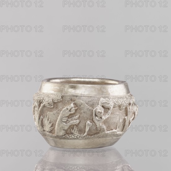 Bowl, South Asian, Indian; applied art vessel 19th/20th century. Depicts hunting iconography: galloping rider on horseback, tigers, horses, elephants, warriors, plants. Weight 155 g; height 5.4 cm; diameter 8.0 cm., A small rounded metal vessel sits against a pale gray background, its surface showing a lustrous silver color with slightly darker toned areas where texture and relief appear. The body of the vessel features a decorative band with raised sculpted scenes in the same silver hue: a few stylized plant forms, a group of animal-like figures with open mouths and raised limbs, and a human-like figure in a dynamic pose. The rim and base are smoother and shinier, while the central decorative area has a more matte, textured appearance. Several leafy motifs and clustered rounded shapes are incorporated into the relief, and the piece rests on a reflective surface that shows a faint silver-toned reflection.