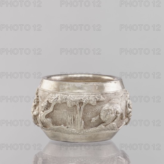 Bowl, 19th/20th century India, South Asian; applied art vessel depicting hunting scenes with tigers, horses, elephants, galloping rider on horseback, warriors, and plants; weight 155 g, height 5.4 cm, diameter 8.0 cm., A small rounded metal vessel with a polished rim and a textured band around its middle decorated in low relief; the relief shows a scene with a tree, a group of organic shapes and figures with flowing forms, and decorative borders above and below the band. The vessel sits on a reflective surface and is shown against a plain background. Visible colors include silver, light gray, and muted metallic tones.