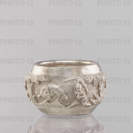 Bowl, South Asian, Indian, applied art, 19th/20th century, India; vessel depicting hunting iconography: tigers, horses, elephants, galloping rider on horseback, warriors, plants; weight 155 g, height 5.4 cm, diameter 8.0 cm., A small round vessel with a metallic silver surface sits against a pale gray background, showing a band of raised sculpted scenes around its midsection with a textured backdrop; the relief includes a figure in motion near a reclining animal, tree-like forms and foliage, and a smooth polished rim and base that reflect light, with overall colors of silver, light gray, and subtle darker gray shadows.