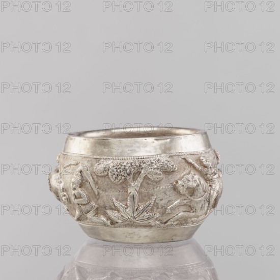 Bowl, South Asian, Indian; applied art vessel, 19th/20th century India; hunting iconography: tigers, horses, elephants, galloping rider on horseback, warriors, plants; weight 155 g, height 5.4 cm, diameter 8.0 cm, A small rounded metal bowl rests on a reflective surface against a pale gray background, the bowl showing a metallic silver tone with areas of slightly darker patina; its exterior is decorated with a raised relief scene that includes stylized trees with textured canopies, leafy plants at the base, and human figures in dynamic poses, all in the same silver color with subtle shadowing that highlights the carved details.
