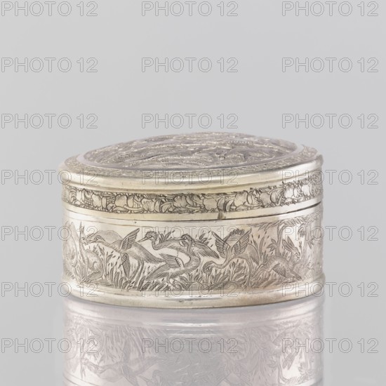 Box with a lid; maker, 19th century, India; Indian; applied art; vessel; materials: tin; decorations: animals—lions, antelopes, jackals, cranes—trees, plant and geometric ornaments; height 3.5 cm, diameter 6.5 cm, weight 105.0 g., A small oval metal box with a fitted lid sits against a pale gray background, the metal showing a silvery tone with subtle darker patina in engraved areas; the lid surface and the sides are decorated with intricate engraved scenes showing a group of birds among grasses and plants, and a decorative floral border runs near the top edge; the box rests on a reflective surface that mirrors the engraved patterns faintly, and visible colors include silver, gray, and darker gray tones.