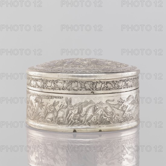 Box with a lid; maker, 19th century India; Indian; materials: tin container; applied art; subject/iconography: animals — lions, antelopes, jackals, cranes; trees, plant ornaments, geometric ornaments; decorations; function: vessel; dimensions: height 3.5 cm, diameter 6.5 cm, weight 105.0 g., A small round metal box with a domed lid sits on a reflective surface; the box is silver in color with intricate engraved decoration all around, including a band of floral or foliate motifs near the top and a lower frieze showing a group of rabbits amid grass and plants and a hint of distant hills, all rendered in fine etched lines; the lid also carries dense engraved patterns, and the reflection of the decorated side and base appears faintly beneath the box against a pale gray background with soft even lighting.