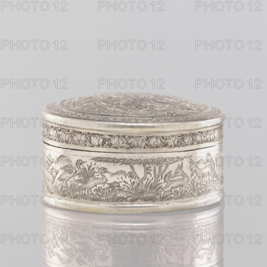 Box with a lid, 19th century, India. Painted metal vessel height 3.5 cm, diameter 6.5 cm, weight 105 g decorated with animals—lions, antelopes, jackals, cranes—trees, plant and geometric ornaments., A small round metal box with a decorated lid sits against a pale gray background, the box and its reflection showing a silver color with engraved scenes around the sides and top; the engravings depict animals and foliage in a darker etched silver tone that contrasts with the smoother silver surface, and a band of floral or leafy ornament wraps near the top edge, while the lid features concentric decorative designs in similar silver hues.