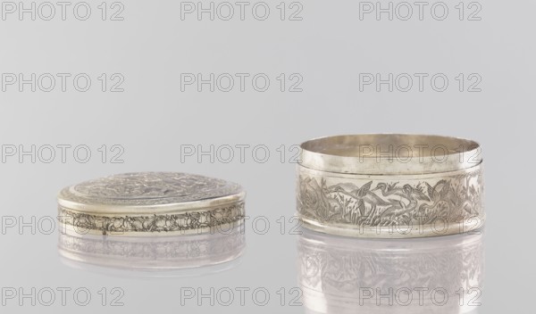 Box with a lid, 19th century, India; Indian; painted metal vessel with applied decoration depicting animals—lions, antelopes, jackals, cranes—trees, plant and geometric ornaments; height 3.5 cm, diameter 6.5 cm, weight 105.0 g., A reflective scene shows a group of two round metal containers placed on a glossy surface, one lying flat with its lid on and the other standing open beside it; both objects have intricate engraved designs around their sides and lids featuring ornate patterns and figurative motifs. The colors visible are silver on the metal surfaces, pale gold tones on some inner and outer edges, a neutral light gray background, and subtle reflections creating softer gray and muted silver hues on the surface beneath the objects.