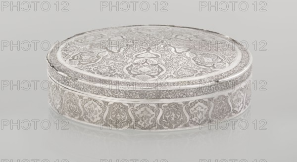 Lidded box, Persian, Qajar dynasty 1785–1925, 19th–20th century. Material: metal vessel. Decoration: geometric and floral motifs, plant ornament. Origin: Isfahan, Iran. Dimensions: height 3.0 cm; diameter 13.0 cm; weight 285 g., A round, flat metal box with an ornate, engraved surface sitting on a reflective pale background; the box displays intricate floral and scrollwork patterns across its top and sides with repeating decorative motifs and bordered bands; visible colors include silver, light gray, and off-white.