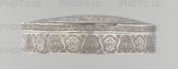 Lidded box, Persian, Qajar dynasty 1785–1925, 19th/20th century, Iran, Isfahan. Brass vessel with geometric and floral motifs plant ornament. Height 3.0 cm; diameter 13.0 cm; weight 285 g., The image shows a decorative oval object with an ornate surface pattern, photographed against a light gray background; the object features a raised domed top and a vertical band around its side with repeating floral and arabesque motifs, and a narrower floral frieze near the top edge; visible colors include shades of silver, white, gray, and pale beige.