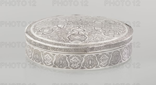 Lidded box, Islamic art, Persian, Qajar dynasty 1785–1925, geometric and floral motifs, plant ornament, 19th–20th century, Iran, Isfahan, vessel, weight 285 g, height 3.0 cm, diameter 13.0 cm, A round, low-profile box with an ornate surface pattern sits against a pale background, showing a predominantly silver and gray color scheme with subtle white highlights; the lid features an intricate floral and curvilinear engraved design covering its entire top, while the side band shows repeating decorative medallions separated by small vertical elements, all rendered in the same silver-gray tones; the rim has a narrower engraved border encircling the lid, and the object casts a soft reflection on the smooth, light-colored surface beneath it.