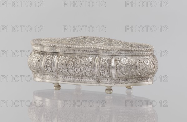 Box with lid, plant ornament, Indian, 19th/20th century, India, height 4.5 cm, width 12.5 cm, depth 6.5 cm, weight 151 g, A small ornate metal box sits on a reflective surface, showing detailed raised floral and scrollwork patterns all over its curved sides and domed lid; the box rests on a few rounded feet and displays an overall metallic silver color with subtle darker patina in recessed areas and hints of warm golden tones on the feet, set against a pale neutral gray background.