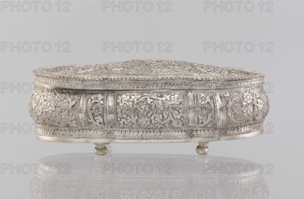 Box with lid, 19th/20th century, India; Indian art; plant ornament; height 4.5 cm, width 12.5 cm, depth 6.5 cm, weight 151 g., A silver decorative box with an oval shape rests on a reflective surface; the box features an intricately embossed floral pattern covering its sides and lid, with repeated circular floral medallions and scrolling vine motifs. The lid is domed and decorated with openwork and textured borders, while the body has alternating raised panels and recessed floral designs. The box stands on three small rounded feet. Visible colors in the image include silver, light gray, a few darker gray tones in the shadows, and a pale neutral background gray.