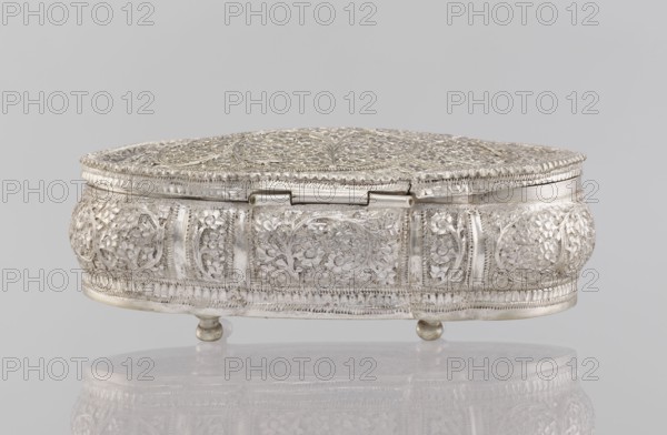 Box with lid; maker, Indian; plant ornament; 19th/20th century, India; height 4.5 cm, width 12.5 cm, depth 6.5 cm; weight 151 g., A decorative metal box with an ornate lid and rounded sides sits on small rounded feet, showing intricate raised floral and vine patterns across its top and sides. The surface appears metallic and reflective, primarily silver in color with subtle variations in tone, and there is a pale gray background and a faint reflection of the box on the surface beneath it.