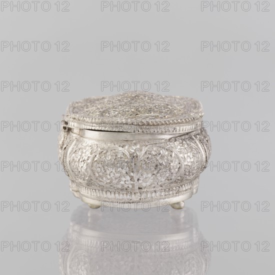 Box with lid, Indian, 19th/20th century, India; plant ornament; height 4.5 cm, width 12.5 cm, depth 6.5 cm, weight 151 g., A small ornate metal box with a domed lid sits on a reflective surface against a plain light gray background; the box is silver in color with intricate raised floral and vine patterns covering its rounded sides and top, a delicate braided edge around the lid, a hinge on one side, and short rounded feet lifting it slightly above the surface, with the silver tones reflected faintly beneath it.