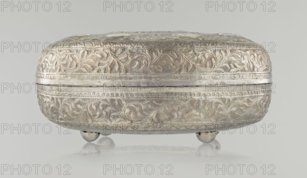 Box with lid; maker, Islamic, Turkish, Ottoman dynasty 1299–1922; 19th century; plant and geometric motifs; materials/technique: varia; dimensions: height 9.0 cm, width 19.5 cm, depth 15.7 cm; weight 210 g., A rounded, ornate metal box with a hinged lid rests on a group of small rounded feet; the surface shows detailed raised floral and vine patterns with textured backgrounds and banded decorative borders. The object appears in silvery and pale gold tones with areas of darker gray patina, set against a light gray background and reflected faintly on a smooth surface beneath it.