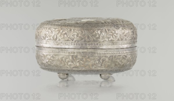 Box with lid, Ottoman, Turkish, 19th century. Wood with plant and geometric motifs; height 9.0 cm, width 19.5 cm, depth 15.7 cm, weight 210 g., A rounded decorative metal container sits on a reflective surface, supported by a few rounded feet; the container has ornate raised floral and foliate patterns in relief encircling its sides and top with beaded borders and horizontal bands; the metal has a silvery appearance with areas of darker patina and subtle warm highlights; the background and surface are pale gray.
