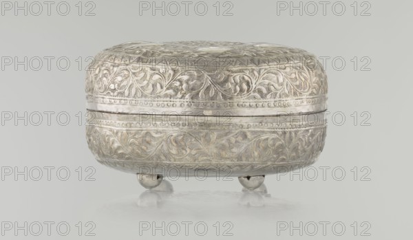 Box with lid, Islamic art, Turkish, Ottoman dynasty 1299–1922, plant motifs, geometric motifs, 19th century, Turkey, varia; height 9.0 cm, width 19.5 cm, depth 15.7 cm, weight 210 g., A rounded decorative metal box with an embossed floral pattern sits on a smooth reflective surface, supported by a few small round feet; the box shows tones of silver with subtle areas of warm beige or light gold, and the background is a pale gray.