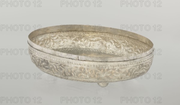 Box with lid, Islamic art, Turkish, Ottoman dynasty 1299–1922, 19th century. Decoration: plant and geometric motifs. Materials/type: box with lid; dimensions: height 9.0 cm, width 19.5 cm, depth 15.7 cm; weight 210 g. Varia., An oval decorative bowl with ornate raised floral and leaf patterns along its exterior and interior surfaces, resting on small rounded feet; the bowl appears metallic with a silvery sheen and areas of warmer, slightly golden patina, set against a pale neutral background in colors of light gray and off-white.