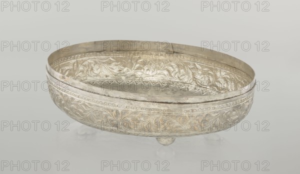 Box with lid; creator, purchase provenance; Islamic art; Turkish; Ottoman dynasty 1299–1922; plant motifs; geometric motifs; 19th century; Turkey; varia; height 9.0 cm; width 19.5 cm; depth 15.7 cm; weight 210 g, A shallow oval metal bowl with ornate raised floral and scrollwork patterns along its outer and inner surfaces, resting on small rounded feet; the bowl appears metallic with visible areas of silver and muted gold tones, along with some darker tarnished gray accents, set against a smooth pale gray background.