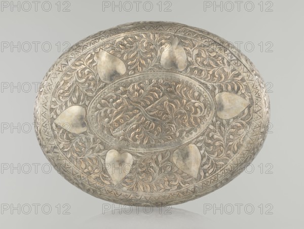 Box with lid, Islamic art, Turkish, Ottoman dynasty 1299–1922, plant motifs, geometric motifs, 19th century, Turkey, height 9.0 cm, width 19.5 cm, depth 15.7 cm, weight 210 g, A round, shallow metallic object with an ornate raised design fills the image; it has a central oval area with swirling vine-like motifs and a branching element, surrounded by a band of repeating raised dots and a border of leafy scrollwork. Around the central band are a few heart-shaped raised forms set within dense foliate patterns, and the outer rim shows a repeated geometric band and additional scrolling leaf decoration. The surface displays a mix of metallic tones including silver, muted gold, and pale gray, with darker gray areas in recessed details and slight warm highlights on raised areas. The background is a smooth, light gray.