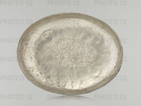 Box with lid, Islamic art, Turkish, Ottoman dynasty 1299–1922, 19th century. Decorated with plant and geometric motifs. Materials unspecified. Dimensions: height 9.0 cm; width 19.5 cm; depth 15.7 cm; weight 210 g., A shallow round metal tray with a slightly raised rim, shown from above, displaying an embossed floral medallion in the center and a decorative border near the edge; the surface has uneven texture and subtle mottling. The visible colors are silvery gray and pale gray with faint darker gray tarnish spots, and a neutral light gray background.