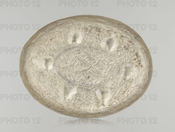 Box with lid, Islamic art, Turkish, Ottoman dynasty 1299–1922, plant motifs, geometric motifs, 19th century, Turkey, varia; height 9.0 cm, width 19.5 cm, depth 15.7 cm, weight 210 g., A shallow round decorative metal tray with an embossed floral and scrollwork pattern across its surface, showing a central oval cartouche surrounded by ornate leaf-like motifs and textured background. The tray displays a few raised, heart-shaped impressions around the inner area and a patterned rim with repeating geometric shapes. The metal has a reflective, metallic sheen with visible variations in tone ranging from bright silver to muted gray and pale gold, and there are areas of darker tarnish and subtle shadowing. The tray rests on a light, neutral background with a faint reflection visible beneath it.
