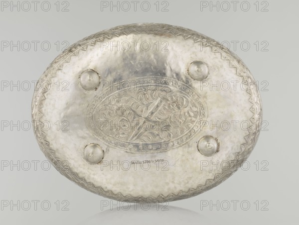 Box with lid, Islamic art, Turkish, Ottoman dynasty 1299–1922, plant motifs, geometric motifs, 19th century, Turkey, varia; height 9.0 cm, width 19.5 cm, depth 15.7 cm, weight 210 g, An oval metal object with a reflective silver surface fills the image, showing an embossed central oval panel with intricate swirling leaf-like patterns and a raised decorative border, surrounded by a smooth area punctuated by a few rounded protrusions; the edge has a repeating triangular zigzag motif, and the overall colors visible are silver, pale gray, and hints of darker gray patina.