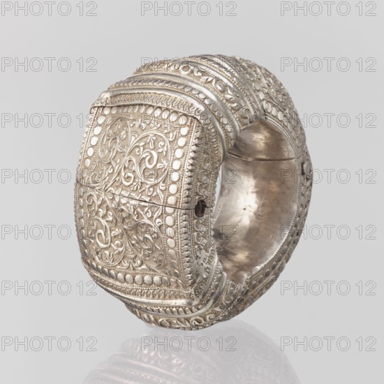 Bracelet, South Asian, Indian. Ring-shaped bracelet decorated with a stylized tendril; rectangular field framed by pearls with tendril. Pin closure. 19th–20th century, India. Jewelry. Weight 460 g; height 11.4 cm; width 8.5 cm; diameter 12.5 cm., A close-up of an ornate metal bracelet with a wide, rounded band and a square, raised central panel covered in intricate filigree and raised beadwork; the surface shows swirling floral and vine motifs in relief within bordered sections, with textured ridges and repeating decorative bands wrapping around the piece. The metal appears silvery with areas of warmer, slightly tarnished patina and subtle variations in sheen. The interior of the bracelet is smooth and reflective with visible wear marks. The image background is a neutral light gray, and the piece casts a faint reflection below it.