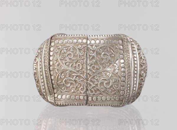 Bracelet, South Asian, Indian. Ring-shaped bracelet decorated with a stylized tendril and a rectangular field framed by pearls with tendril; pin closure. 19th–20th century, India. Jewelry. Weight 460 g; height 11.4 cm; width 8.5 cm; diameter 12.5 cm., A small ornate metal box with a hinged center, shown closed and resting on a reflective surface; the box has an overall silver color with areas of darker patina, rounded ends, and a rectangular central panel on top featuring a symmetrical raised floral and vine pattern surrounded by a border of raised circular dots and decorative beaded and zigzag edges, with similar floral and ornamental detailing continuing onto the curved sides.