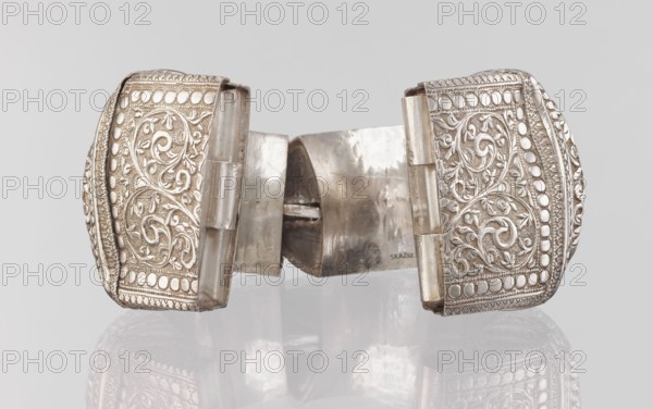 Bracelet, South Asian, Indian. Ring-shaped bracelet decorated with a stylized tendril; rectangular field framed by pearls with tendril. Pin closure. 19th–20th century, India. Jewelry. Weight 460 g; height 11.4 cm; width 8.5 cm; diameter 12.5 cm., A metallic bracelet with an open cuff shape sits against a light grey background, showing a reflective surface underneath. The bracelet is silver in color with ornate embossed panels on each end featuring swirling vine and leaf patterns framed by rows of raised rounded shapes and textured borders. The central band connecting the two ornate ends is smoother and slightly worn, showing reflective highlights and a few subtle scratches. The overall tones visible are silver, grey, and off-white reflections, with darker grey areas in the shadowed recesses and a faint mirrored reflection of the piece below.