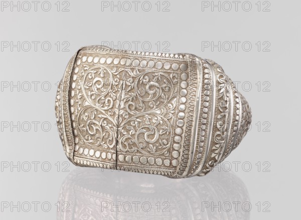 Bracelet, South Asian, Indian. Ring-shaped bracelet decorated with a stylized tendril, featuring a rectangular field framed by pearls with tendril. Pin closure. 19th–20th century. Materials: plant ornament note: jewelry. Weight 460 g; height 11.4 cm; width 8.5 cm; diameter 12.5 cm. Geometric ornaments, decorations., A detailed metallic object with an ornate surface featuring intricate raised floral and vine patterns and repeating circular motifs, shown against a plain light gray background; the object appears reflective with a polished silver tone and areas of darker patina, and its surface shows carved textures and bands of decorative ridges, with a soft reflection visible beneath it.