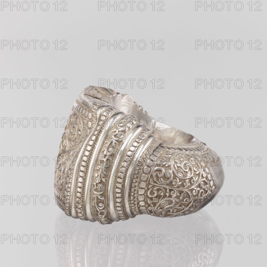 Bracelet, Ring-shaped bracelet decorated with a stylized tendril; rectangular field framed by pearls with tendril. Pin closure. 19th–20th century, India. Materials: plant ornament? jewelry. Weight 460 g; height 11.4 cm; width 8.5 cm; diameter 12.5 cm. South Asian/Indian cultural origin; geometric ornaments, decorations., A close-up view of an ornate metallic ring with intricate raised filigree and beaded patterns in silver tones, showing swirling floral and leaf motifs and several smooth ridged bands; the background is a pale light gray and the ring casts a soft reflection on the surface below.