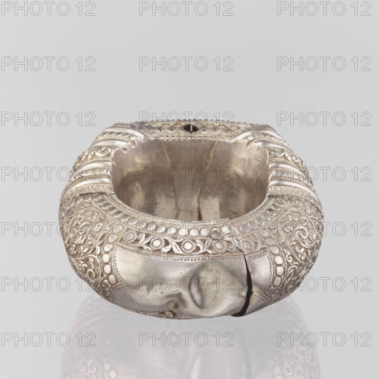 Bracelet, South Asian, Indian. Ring-shaped bracelet decorated with a stylized tendril; rectangular field framed by pearls with tendril; pin closure. 19th–20th century, India. Jewelry. Weight 460 g; height 11.4 cm; width 8.5 cm; diameter 12.5 cm., A rounded metal vessel with a hollow center sits against a pale gray background, its surface showing ornate raised patterns of scrolling floral and circular motifs around the exterior. The metal appears silver in color with areas of darker tarnish and patina, and the interior shows a smoother, slightly duller silver tone. A visible crack runs from the rim down the side, and the rim itself is scalloped with decorative beadwork. The piece casts a faint reflection on the glossy surface beneath it.
