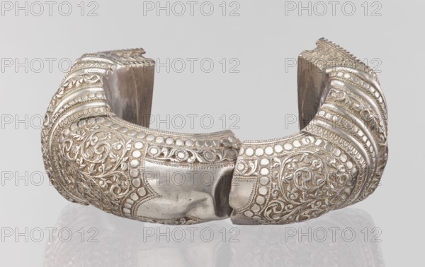 Bracelet, South Asian, Indian; ring-shaped bracelet decorated with a stylized tendril and a rectangular field framed by pearls with tendril; pin closure. 19th–20th century, India; jewelry; weight 460 g; height 11.4 cm; width 8.5 cm; diameter 12.5 cm., A curved metallic cuff with ornate raised floral and scrollwork patterns, featuring rows of rounded bead-like elements and embossed ridges, shown against a light gray background; the cuff displays a polished silver tone with areas of darker patina and subtle brownish discoloration, and its reflective surface casts a faint mirror image on the pale surface beneath.