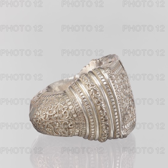 Bracelet, South Asian, Indian. Ring-shaped bracelet decorated with a stylized tendril; rectangular field framed by pearls with tendril; pin closure. 19th–20th century, India. Jewelry. Weight 460 g; height 11.4 cm; width 8.5 cm; diameter 12.5 cm., A close-up of a decorative metal cuff with intricate raised patterns, shown against a light gray background; the cuff displays ornate scrolling floral and leaf motifs, bands of rounded raised dots, and several parallel ridged bands with detailed embossing; the surface appears metallic with silvery tones and subtle darker gray shadows in the recesses, and the object casts a soft reflection on the pale surface beneath it.