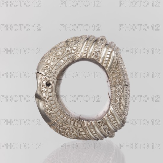 Bracelet, Ring-shaped bracelet decorated with a stylized tendril within a rectangular field framed by pearls and tendril. Pin closure. 19th–20th century, India. Materials: plant ornament note: ornament motif?, jewelry, geometric ornaments, decorations. Weight 460 g; height 11.4 cm; width 8.5 cm; diameter 12.5 cm., A circular ornamental object with an open center, primarily metallic silver in color with areas of pale pink visible on the inner edge, featuring ornate raised patterns including swirls, floral motifs, and rows of rounded studs; the surface shows alternating smooth ridges and intricately textured bands, and a small dark circular hole is visible on one side; the object rests against a light gray background and casts a faint reflection below, with overall colors of silver, pale pink, dark gray, and light gray.