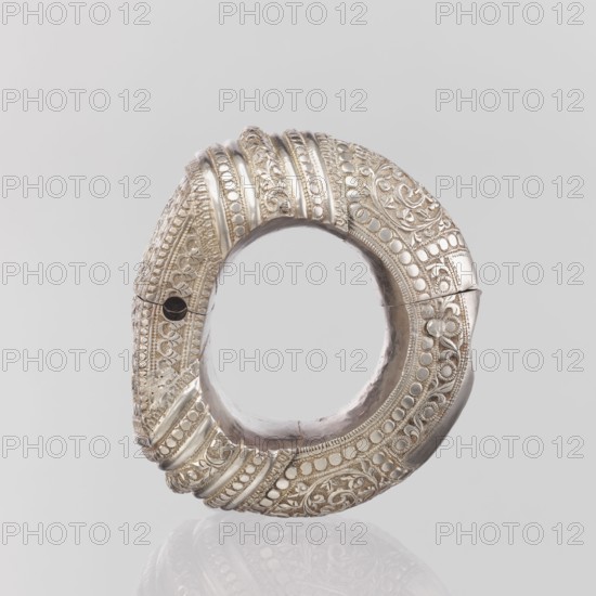 Bracelet, Ring-shaped bracelet decorated with a stylized tendril and a rectangular field framed by pearls with tendril; pin closure. 19th–20th century, India. Materials: plant ornament? uncertain. Style: geometric ornaments, decorations. Jewelry. Weight 460 g; height 11.4 cm; width 8.5 cm; diameter 12.5 cm., A circular metallic object with an open center, appearing in silver and pale gold tones, featuring ornate raised patterns and embossed decorative motifs around its outer ring; the surface shows a mix of smooth and textured areas with rows of rounded beads, floral-like scrollwork, and parallel ridges, and there is a small dark circular hole on one side; the object sits against a light gray background and casts a faint reflection below.