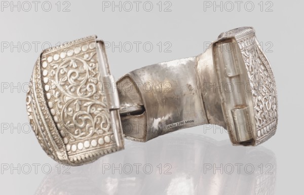 Bracelet, South Asian Indian 19th–20th century. Ring-shaped bracelet of metal jewelry, decorated with a stylized tendril within a rectangular field framed by pearls and tendril motif; pin closure. Dimensions: height 11.4 cm, width 8.5 cm, diameter 12.5 cm; weight 460 g., plant ornament, geometric ornaments., A close-up view of a metallic hinged object with ornate embossed patterns on its two outer panels, showing swirling floral motifs and rows of raised round dots, with a worn central interior surface that is reflective and slightly tarnished; colors visible include silver, gray, off-white, and subtle brownish tones on the metal, set against a pale gray background with a faint reflection of the object below.