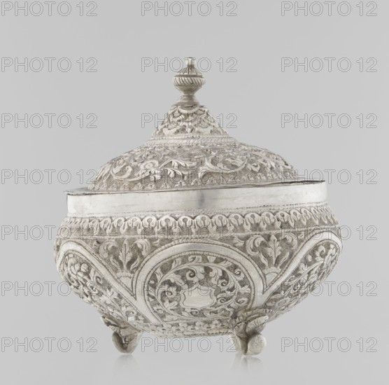 Vessel with a lid, Islamic art, Turkish, Ottoman dynasty 1299–1922, plant ornament with flowers and plant tendril, marked coat of arms, 19th century, Turkey, vessel, weight 240 g, height 11.7 cm, diameter 10.5 cm, An ornate rounded container with a domed lid and a finial on top, each surface covered in raised floral and scrollwork patterns; the body sits on three small rounded feet and has repeating arched medallion designs around its sides, with detailed textured backgrounds and smooth bands near the rim; the visible colors are shades of silver, light gray, and off-white against a pale gray background.