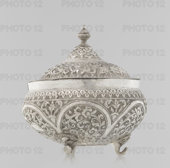 Vessel with a lid, Islamic art, Turkish culture, Ottoman dynasty 1299–1922, plant ornament, flowers, plant tendril, marked coat of arms, 19th century, Turkey, vessel, weight 240 g, height 11.7 cm, diameter 10.5 cm, A decorative lidded vessel with ornate raised floral and scrollwork patterns covering its rounded body and conical lid, resting on three small curved feet; the surface appears metallic with tones of silver, gray, and off-white against a pale gray background.