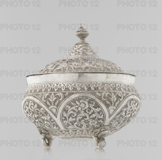 Vessel with a lid, Islamic art, Turkish culture, Ottoman dynasty 1299–1922, plant ornament, flowers, plant tendril, marked coat of arms, 19th century, Turkey, vessel, weight 240 g, height 11.7 cm, diameter 10.5 cm, An ornate lidded vessel with a rounded body and a domed lid topped by a finial, all covered in intricate raised floral and scrollwork patterns; the vessel rests on a few small curved feet and shows a mix of smooth and textured surfaces. The visible colors are silver and gray.