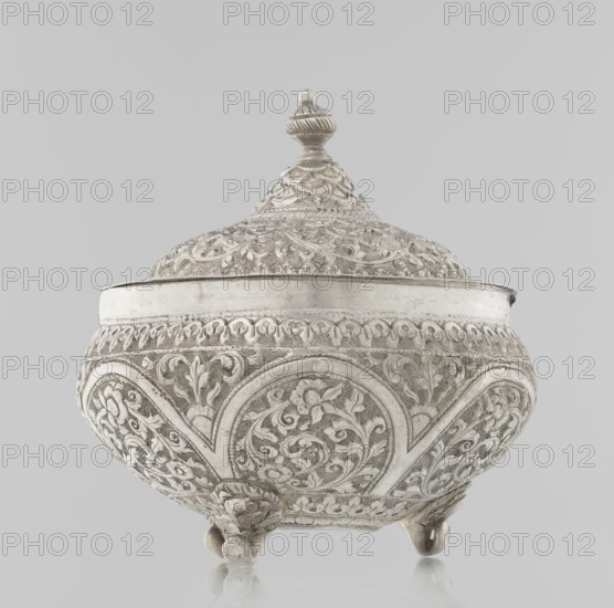 Vessel with a lid, Islamic art, Turkish culture, Ottoman dynasty 1299–1922, plant ornament with flowers and plant tendril, marked coat of arms, 19th century, Turkey, vessel, weight 240 g, height 11.7 cm, diameter 10.5 cm, A decorative lidded bowl or container with an ornate domed lid topped by a finial sits against a plain light gray background. The body and lid display dense, carved or embossed floral and foliate patterns in a metallic surface that appears silver or off-white, with areas of darker patina accenting the relief. The bowl rests on a few rounded feet and shows curved panels of repeating scrollwork and stylized leaves all around. Overall visible colors are silver, off-white, light gray, and touches of darker gray in the recessed details.