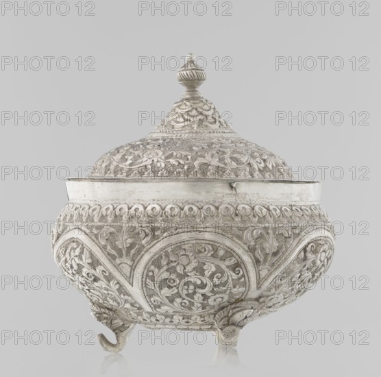 Vessel with a lid, Islamic art, Turkish, Ottoman dynasty 1299–1922, plant ornament, flowers, plant tendril, marked coat of arms, 19th century, Turkey, vessel, weight 240 g, height 11.7 cm, diameter 10.5 cm, A decorative lidded vessel sits against a plain background, showing ornate carved relief all over its rounded body and domed lid; the surface displays intricate swirling floral and foliate patterns, repeated circular medallion motifs, and a band of repeating scallop-like details near the rim, while the lid is topped with a finial that has a fluted and rounded shape. The vessel rests on a few short curved feet and has a gently flared rim. The visible colors are shades of silver, gray, and off-white, with subtle variations creating highlights and shadows across the carved surface.