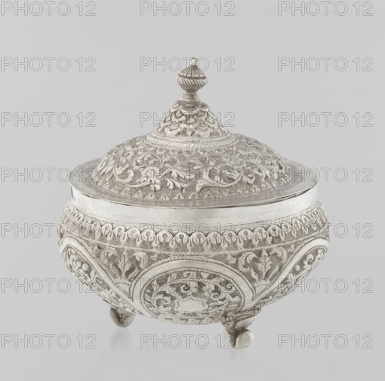 Vessel with a lid, Islamic art, Turkish culture, Ottoman dynasty 1299–1922, plant ornament, flowers, plant tendril, marked coat of arms, 19th century, Turkey, vessel, weight 240 g, height 11.7 cm, diameter 10.5 cm, A round ornate container with a domed lid topped by a finial, all featuring raised decorative patterns of scrolling foliage and floral motifs; the body rests on a few small rounded feet and shows repeating arched panels with intricate relief work. The visible colors are silver and shades of grey against a light grey background.