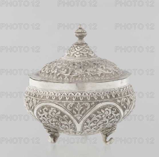Vessel with a lid; creator, 19th century; Turkish, Ottoman dynasty 1299–1922; Islamic art; plant ornament with flowers and plant tendrils; marked with a coat of arms; vessel; weight 240 g; height 11.7 cm; diameter 10.5 cm., A decorative lidded container with ornate raised floral and scrollwork patterns across its rounded body and domed lid, topped with a small finial; the piece stands on a few short curved feet and features detailed carved motifs and textured surfaces. Colors visible include silver, gray, and off-white.