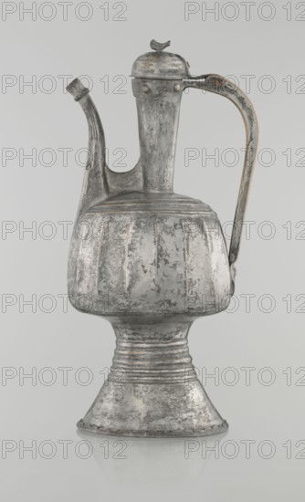 Jug aftabe, Islamic art; 19th century, North Caucasus; water jug aftabe vessel; height 30.0 cm, length 14.8 cm, diameter 12.0 cm., A tall metallic vessel with a rounded bulbous body, a flared pedestal base, a long curved spout, and an arched handle with a hinged lid topped by a small bird-shaped finial; the surface shows worn, textured patches and irregular vertical fluting on the body, with visible seams and rivets where components join, and a reflective sheen creating lighter and darker areas. The visible colors are shades of silver and gray with traces of darker gray patina and faint warm brownish accents along some edges.