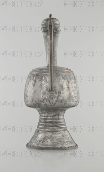 Jug aftabe, 19th century North Caucasus; Islamic art; aftabe water jug; vessel; height 30.0 cm, length 14.8 cm, diameter 12.0 cm, A single metallic vessel with a tall narrow neck rising from a rounded body and flared base is centered against a light gray background; the surface of the object shows a worn, textured finish in shades of silver and gray with darker patches and subtle hints of brownish tarnish; a straight, vertical handle runs from the top of the neck down the front of the body and appears slightly darker with thin lines of brown accent; the rounded body has vertical fluted ridges and a small decorative shield-shaped element where the handle meets the body; the flared pedestal base features concentric horizontal bands and a scalloped edge, and the entire piece is reflected faintly on the smooth surface beneath it.