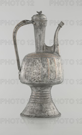 Jug aftabe, unknown provenance; Islamic art; 19th century; North Caucasus; jug/aftabe for water; vessel; height 30.0 cm, length 14.8 cm, diameter 12.0 cm, A metal pitcher with a tall narrow neck, a curved spout, and a high arched handle resting against the side, standing on a flared pedestal base; the surface shows worn, textured patina with areas of roughness and faint vertical ridges around the bulbous body, and a domed hinged lid topped by a small decorative finial; colors visible include shades of gray, silver, and hints of rusty brown.