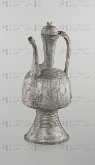 Jug aftabe, 19th century, North Caucasus; Islamic art; material: vessel for water; dimensions: height 30.0 cm, length 14.8 cm, diameter 12.0 cm., The image shows an aged metal ewer with a bulbous body on a flared pedestal base, a tall neck with a hinged domed lid topped by a small decorative finial, a curved handle attached to the neck and body, and a slender spout extending from the shoulder; the surface displays worn areas with a textured, mottled appearance and visible patina in shades of gray, silver, and hints of brownish rust against a plain pale gray background.