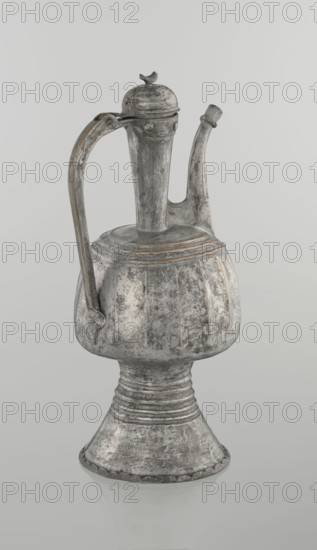 Jug aftabe, unknown provenance; Islamic art; aftabe water jug; 19th century, North Caucasus; vessel; height 30.0 cm, length 14.8 cm, diameter 12.0 cm., A metal ewer with a tall narrow neck, a curved spout, and a high arched handle sits against a plain light gray background; the vessel has a rounded, bulbous midsection above a flared, ridged foot and a hinged lid topped with a small bird-shaped finial. The surface shows a weathered, textured patina with visible areas of wear and scraping, displaying shades of silver-gray with darker gray accents and touches of muted brownish or coppery tones along edges and ridges. The handle and spout have slight discoloration and surface irregularities, and the overall form features subtle ridges and vertical paneling on the body, with a soft reflection visible beneath the base.
