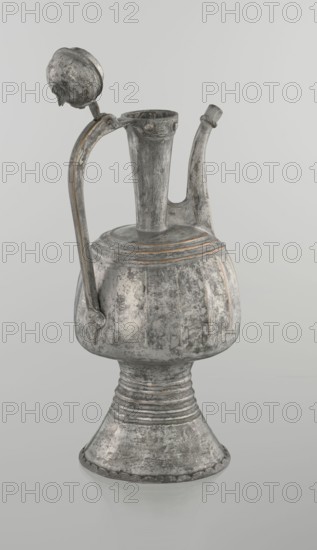 Jug aftabe, Islamic art, jugs, aftabe, water, 19th century, North Caucasus, vessel; height 30.0 cm, length 14.8 cm, diameter 12.0 cm, A metal pitcher with a rounded body, narrow neck, curved spout, and tall flared base sits against a pale background; the surface shows a weathered, textured finish with areas of darker gray, lighter gray, and hints of muted brownish tones; a curved handle rises from the body to the neck and ends near a hinged, domed lid that is open, the lid sharing the same worn gray and brown coloration; the body has subtle vertical ridges and horizontal bands near the top, and the flared pedestal base features concentric ridges and a scalloped edge, all reflecting the same aged gray and brown color palette.